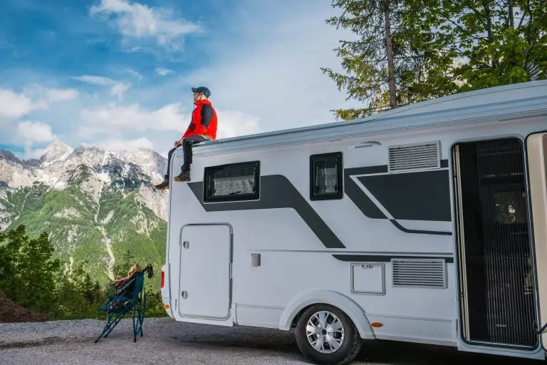 A person in a bright jacket sits on the roof of an RV, taking in the breathtaking view of snow-capped mountains. Nearby, a camping chair is positioned, suggesting a peaceful outdoor experience. Lush greenery surrounds the area, highlighting the natural beauty of the landscape.